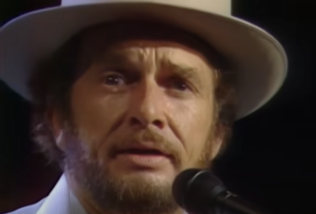 A man with a beard and mustache is wearing a wide-brimmed hat and a light-colored shirt. He is singing into a microphone, with a focused and emotional expression on his face. The background is dark, emphasizing the subject's face and hat.