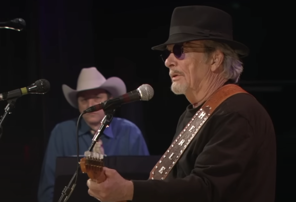 An older man wearing a black hat and sunglasses plays an electric guitar and sings into a microphone on stage. Another man wearing a white cowboy hat stands behind him in the background, also facing a microphone. The setting appears to be a concert or performance.