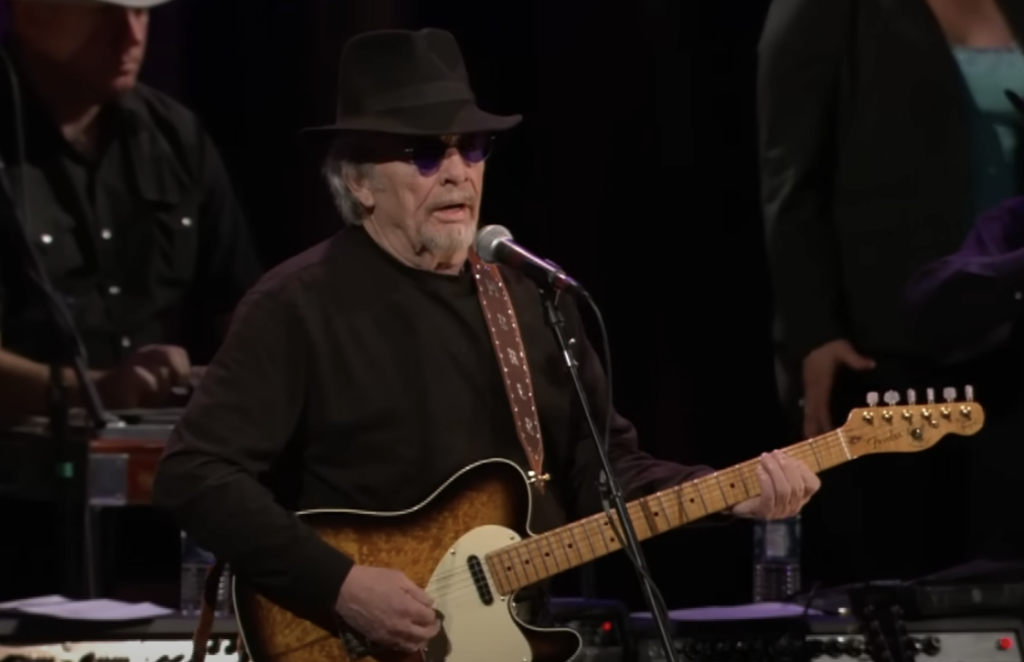 An older man wearing a black hat, dark sunglasses, and a black shirt is playing an electric guitar and singing into a microphone on stage. Two other musicians are partially visible in the background.