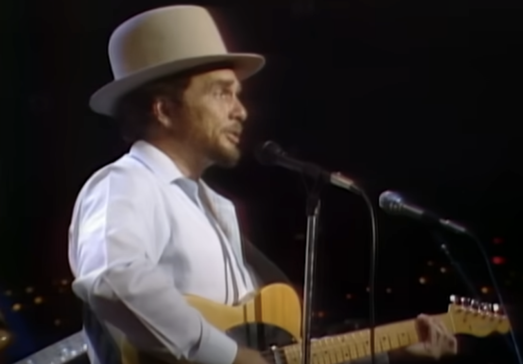 A man wearing a large light-colored hat and a light-blue shirt is singing into a microphone on a stage. He is holding a beige electric guitar. The background is dark with blurry lights, suggesting a nighttime performance.