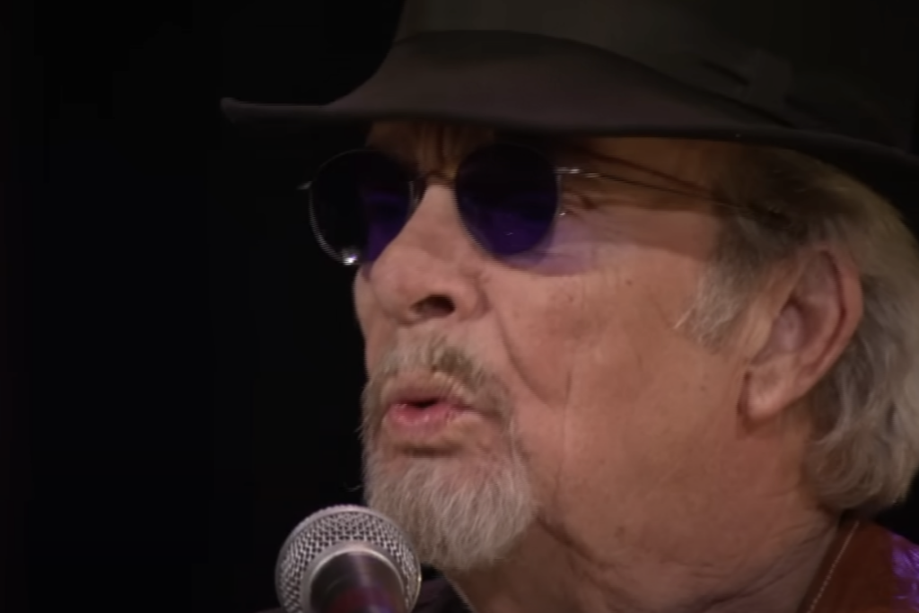 An older man with light hair and a mustache is wearing a black hat, dark sunglasses, and a goatee. He is singing into a microphone against a dark background.