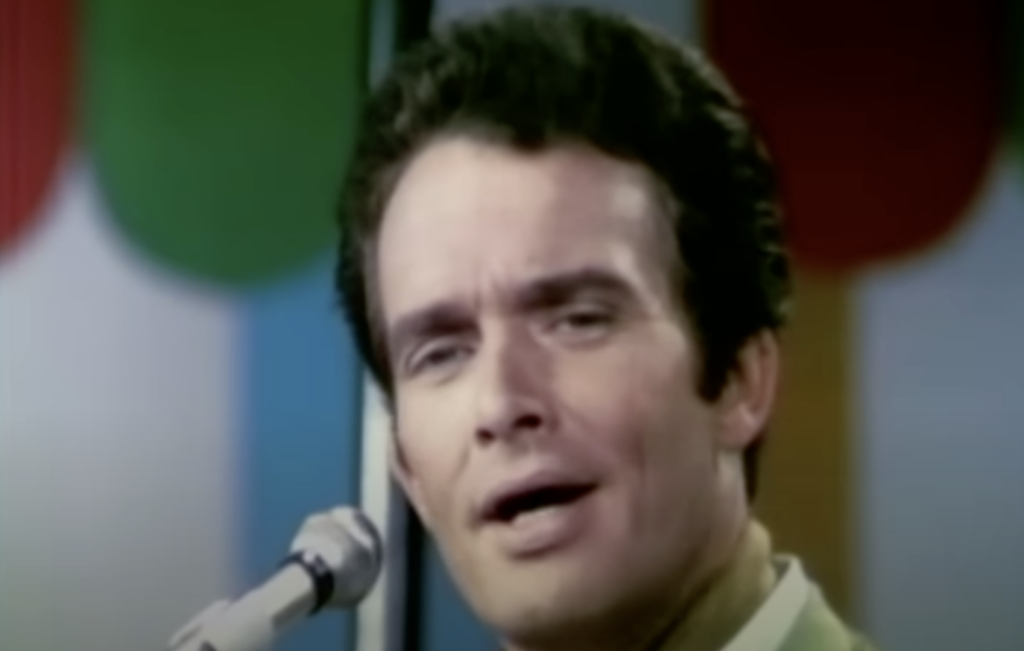 A person with dark hair sings into a microphone. The background is colorful with green, red, blue, orange, and yellow vertical shapes. The person wears a light-colored shirt or jacket and has an expressive facial expression while performing.