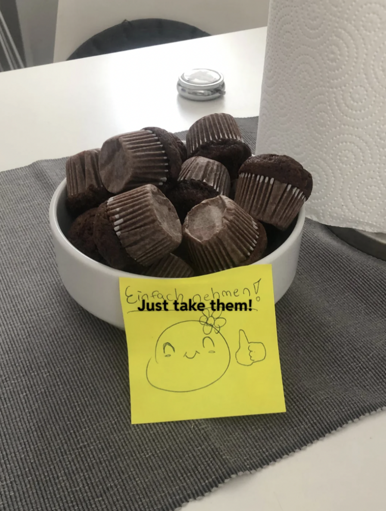 A white bowl filled with chocolate muffins is placed on a gray tablecloth. A yellow sticky note with a smiling face drawing, a flower, thumb-up sign, and text "Einfach nehmen! Just take them!" is attached to the bowl. A roll of paper towels is in the background.