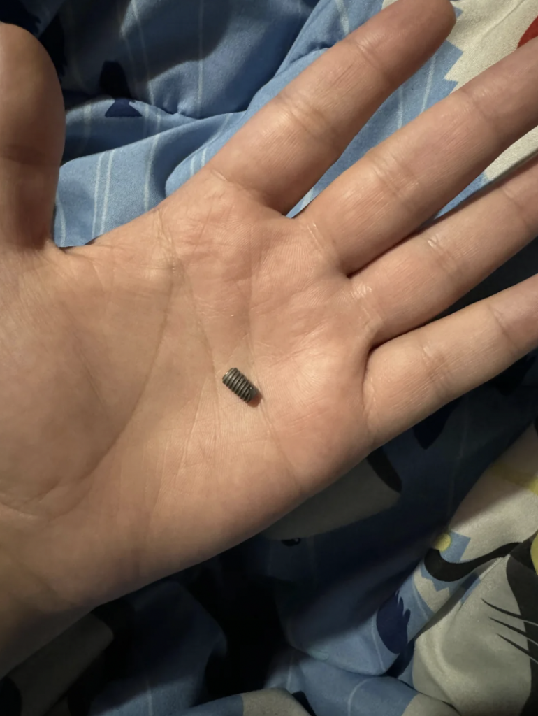 A small metal screw rests on the palm of a person's hand. The background consists of blue and white fabric with various patterns.