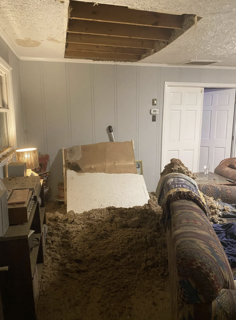 A living room with a large hole in the ceiling, revealing attic insulation debris that has fallen onto the floor and a couch. Pieces of drywall and insulation cover the nearby furniture and floor. Light filters in from a lamp on a side table, illuminating the mess.