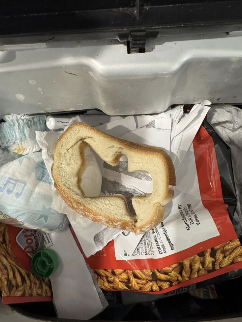 A hollowed-out slice of bread rests atop scattered trash, including a crumpled diaper, plastic bottle cap, and various papers inside an open garbage can.