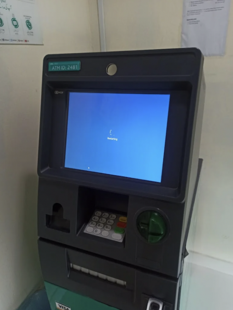 An ATM machine with a large screen displaying a message that says "Restarting." Below the screen are a keypad, card slot, and cash dispenser. There are some papers pinned to the wall in the background. The ATM has an ID label marked "ATM ID: 2481.