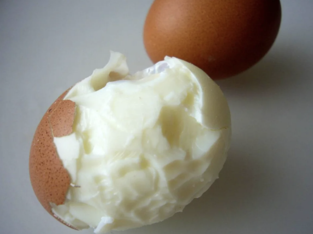 Two brown eggs are shown, one of which is partially peeled, revealing a white interior. Additionally, the peeled egg appears to have a bite taken out of it, exposing more of the white interior and some of the underlying egg structure.