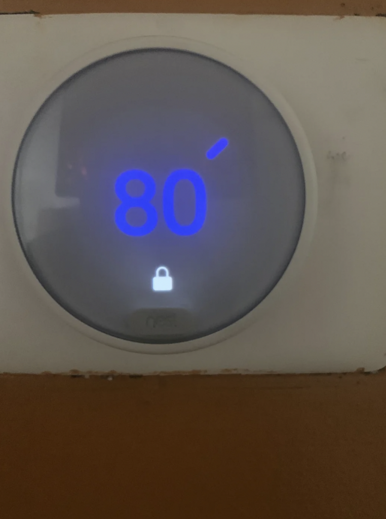 A circular digital thermostat mounted on a wall displays a temperature reading of 80 degrees Fahrenheit in bright blue numbers. A small lock icon is visible below the temperature, indicating that the thermostat settings are locked.