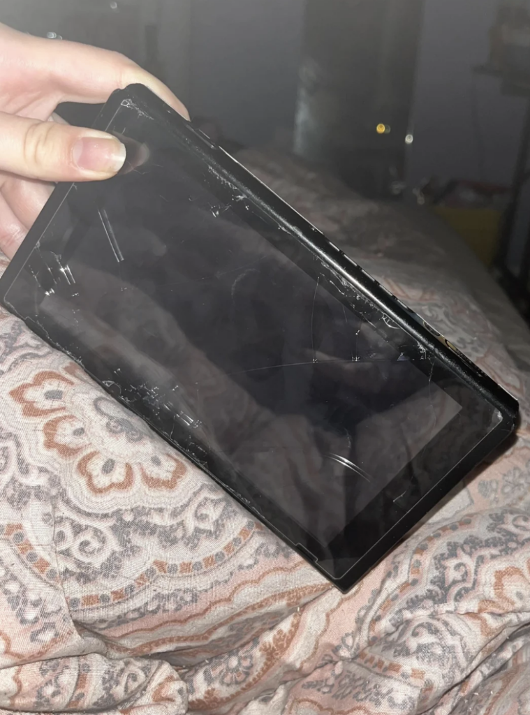 A person holding a cracked and damaged handheld gaming console over a patterned blanket. The screen has several visible cracks and appears broken.