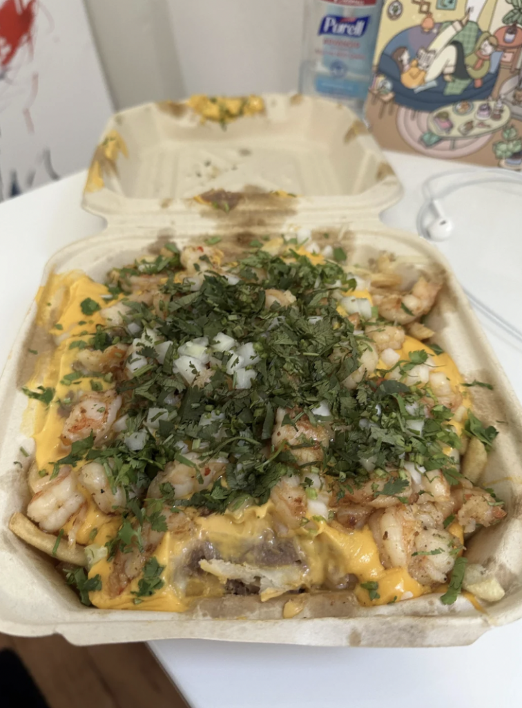 A takeout container filled with loaded fries topped with shrimp, melted cheese, chopped onions, and cilantro sits on a white surface. In the background, a bottle of hand sanitizer and some colorful artwork are partially visible.