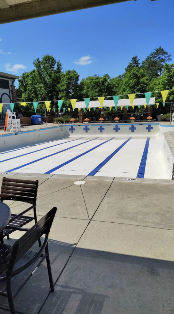 A large, empty outdoor swimming pool with lane markers and cross symbols on the pool floor. There are colorful triangular flags strung above the pool. In the foreground, there are patio chairs and a table on a concrete deck. Trees and buildings are in the background.