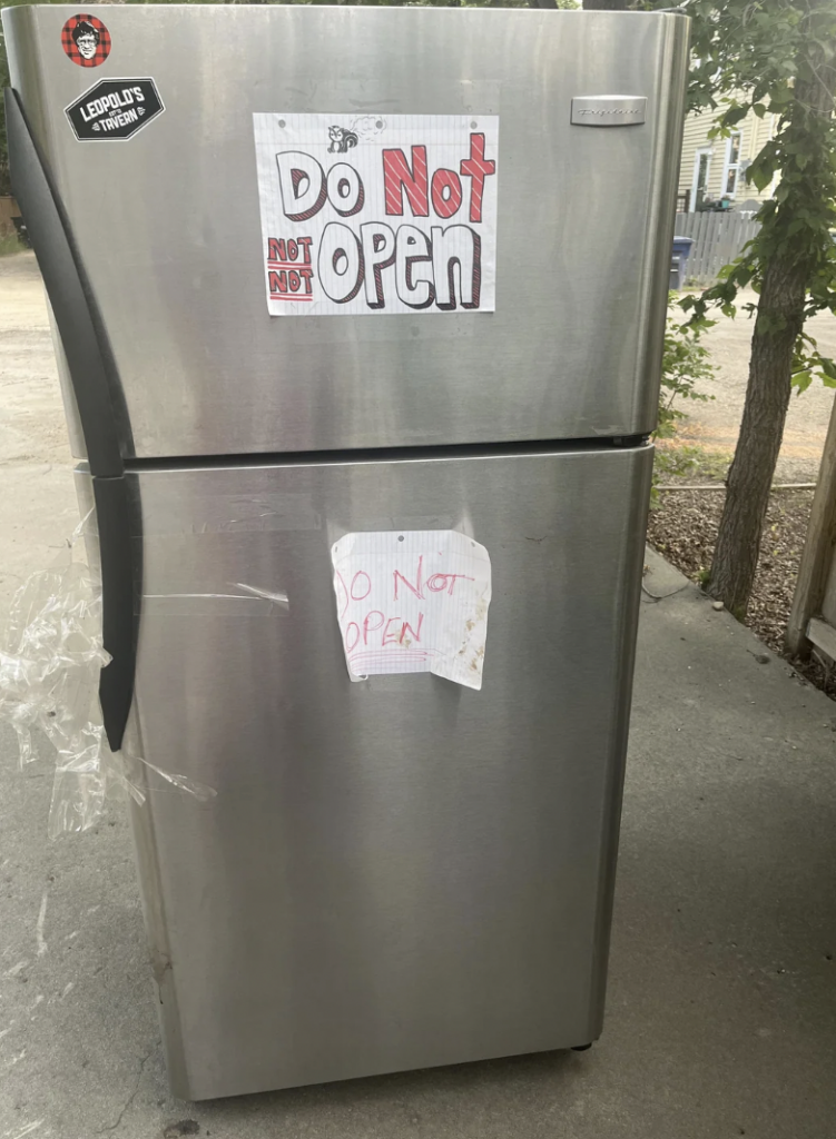 A stainless steel refrigerator is placed outdoors, with two handwritten signs taped to it. The larger sign at the top says "Do Not Open" in bold red and black letters, and the smaller, faded sign below also reads "Do Not Open." Duct tape is visible on the door.