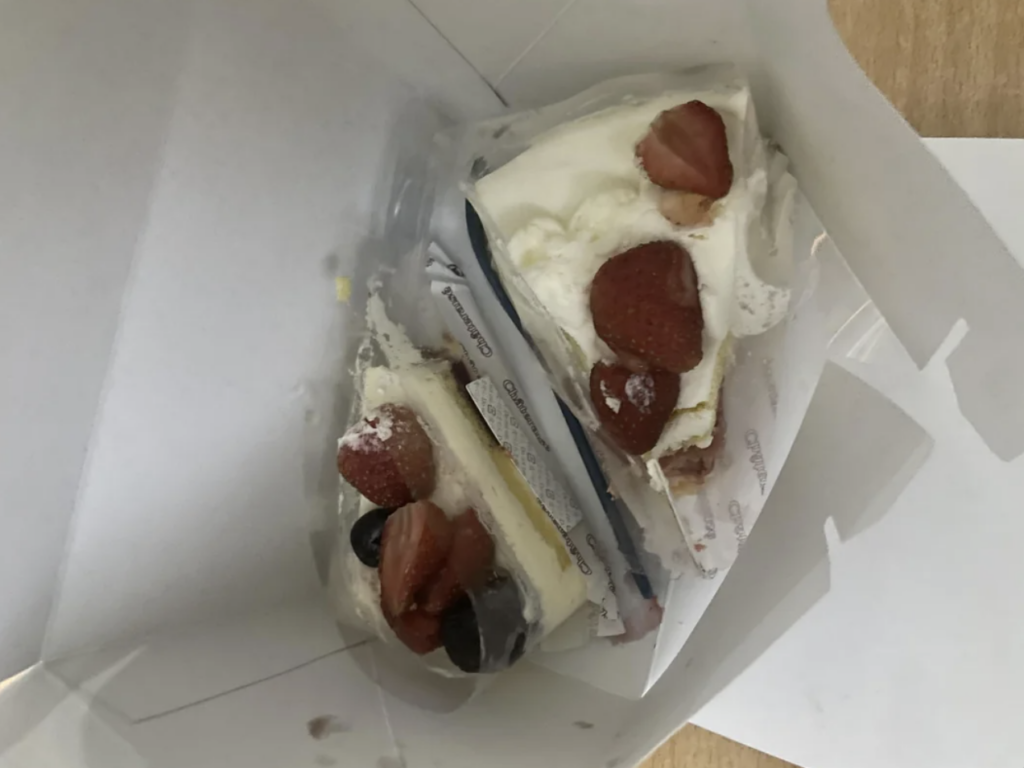 Two slices of cake in a white box. The cakes are topped with whipped cream, strawberries, and blueberries. The box is slightly open, revealing the cakes wrapped in clear packaging. The background is a light-colored wooden surface.