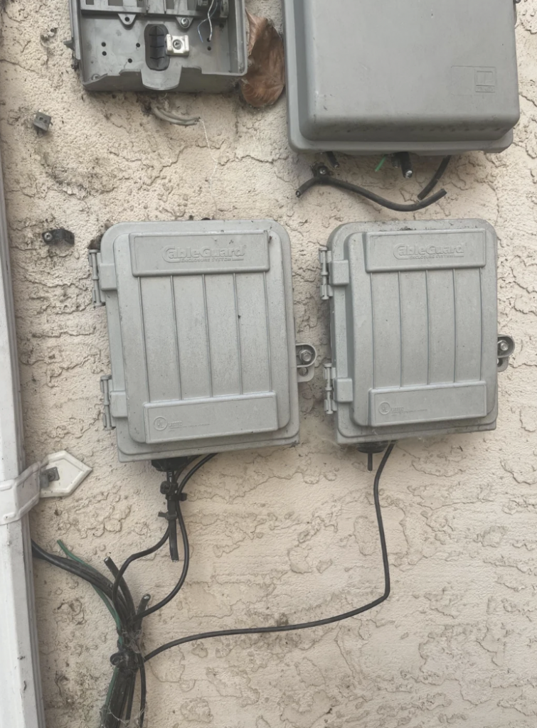 An image showing three outdoor communication utility boxes mounted on a textured cream-colored wall. Two gray rectangular boxes are positioned side by side at the lower part, with wires coming from underneath them. An additional gray box is situated above them.