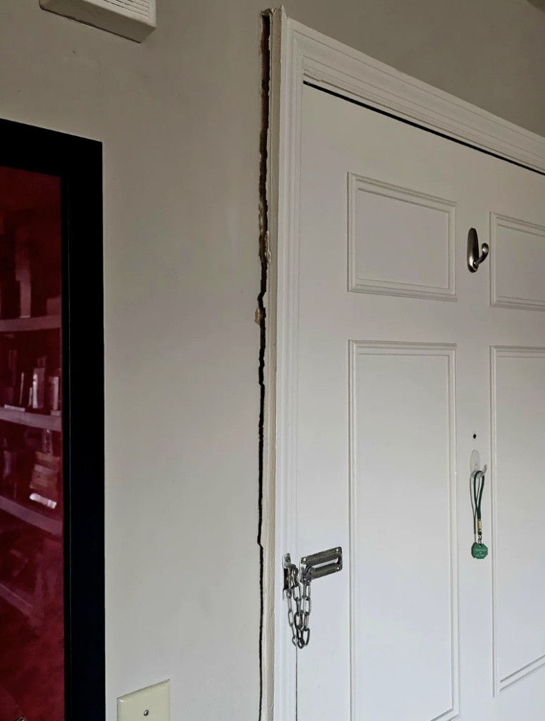 A large crack runs vertically along the wall beside a white interior door, which has a metal chain lock. The crack reveals underlying material. The wall is painted light beige, and there is a red-framed picture partially visible on the left.