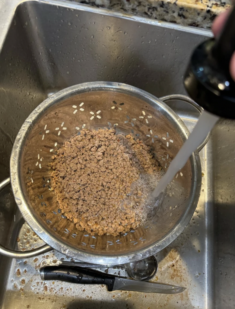 Ground meat is being rinsed under running water in a metal colander positioned over a sink. The sink has a knife, a pair of scissors, and some food residue at the bottom. The faucet is turned on, and water is being sprayed over the meat.