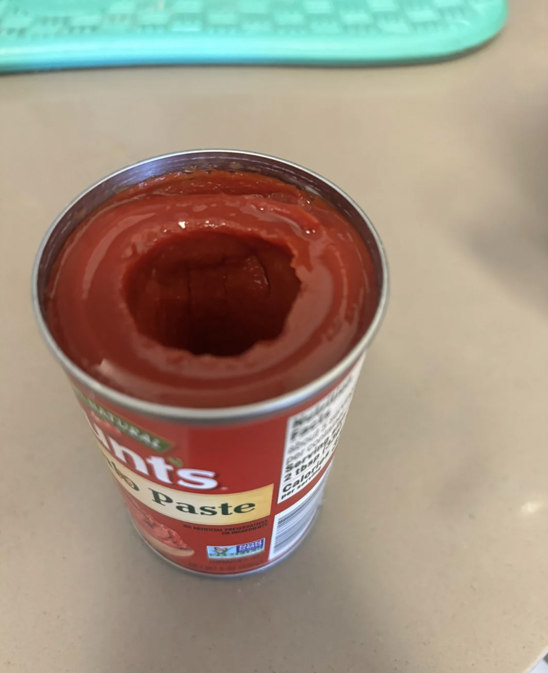 An opened can of tomato paste with a hole in the center, exposing the tomato paste inside. The can's label shows the brand and some text, partially visible. The background appears to include a light-colored surface and a teal object out of focus.