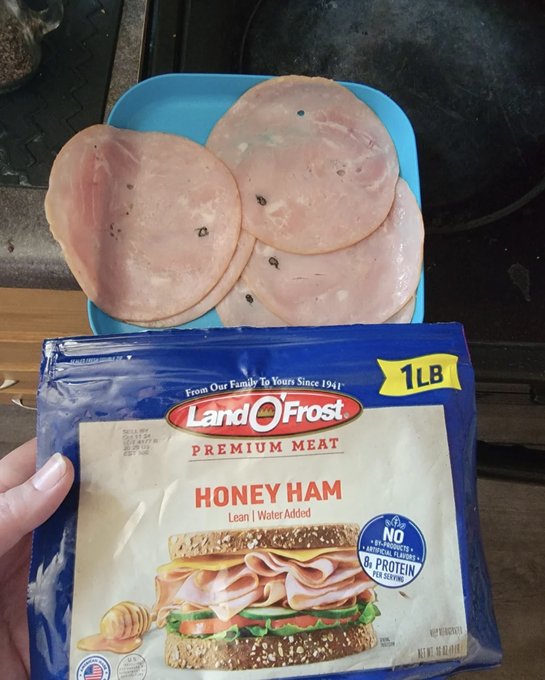 An open package of Land O'Frost Premium Meat Honey Ham is displayed, showing the label that indicates it is a 1-pound package. In the background, several slices of ham are placed on a blue tray. The scene is set on a kitchen counter and stove.