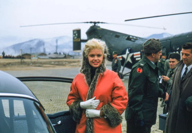 A woman in a bright red coat with a fur collar smiles, standing beside an open car door. Behind her, men in military uniforms gather near a helicopter on a tarmac. Snow-covered mountains are visible in the background.