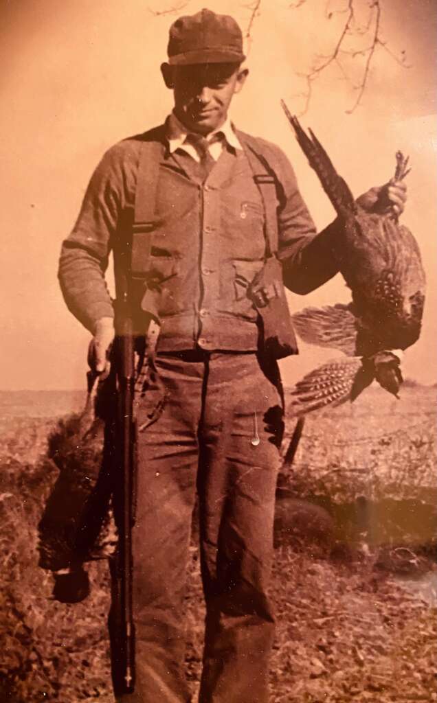 A sepia-toned photograph of a man standing outdoors, holding a rifle in his left hand and several pheasants in his right hand. He is wearing a cap, a buttoned sweater, and dark trousers with suspenders. The background shows a grassy field and trees.