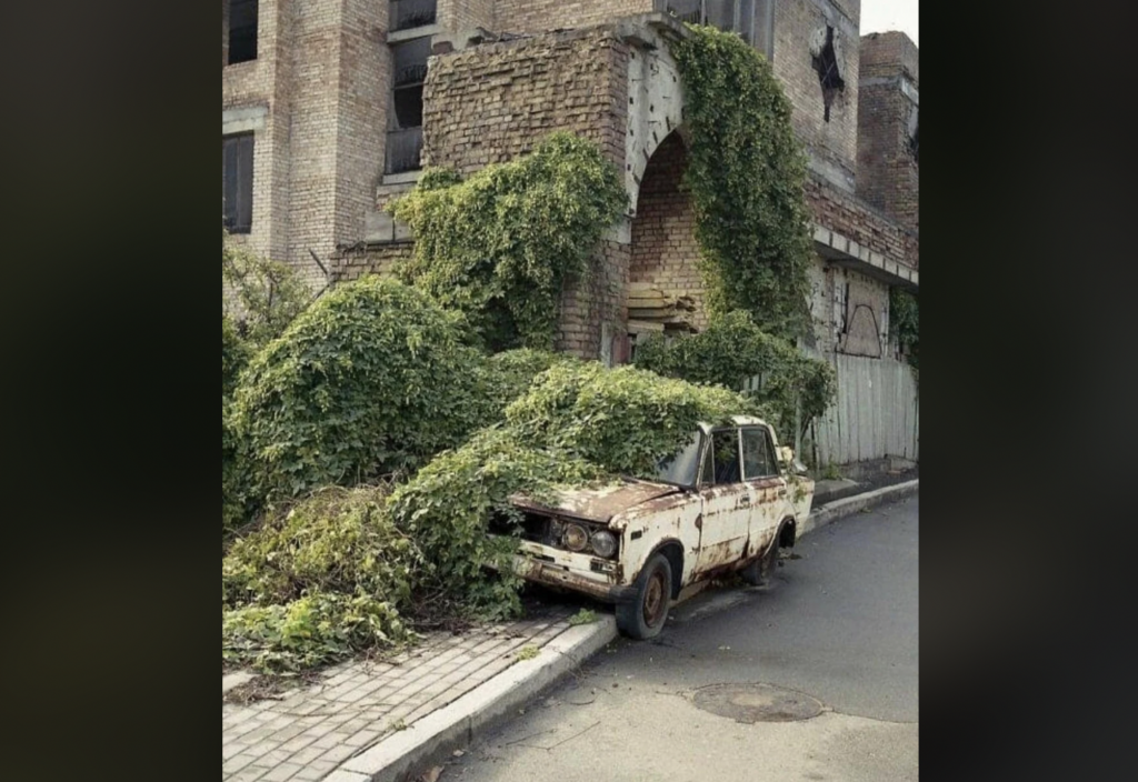 An old, rusted car covered in overgrown ivy is parked on the side of a street in front of an abandoned and decaying brick building. Vegetation has engulfed both the car and parts of the building, giving the scene a sense of abandonment and neglect.