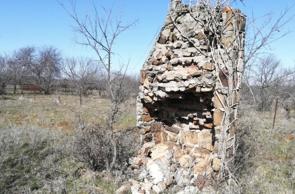 A weathered, partially collapsed stone chimney stands amid a dry, leafless landscape with sparse vegetation and bare trees under a clear blue sky. Vines are seen growing over the structure.