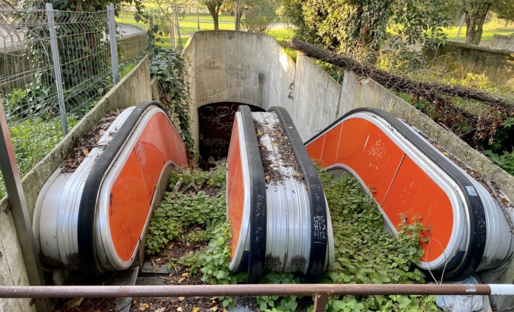 An overgrown, abandoned set of outdoor escalators, flanked by concrete walls, leading down into a dark entrance. Vegetation is growing through the steps and around the sides, indicating long-term disuse. The escalators are lined with faded orange panels.