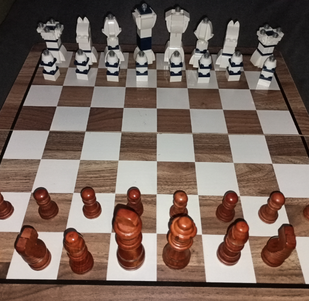 A chessboard is set up for a game, with white pieces made of abstract blocky shapes positioned at the top and traditional brown pieces at the bottom. The board features alternating dark and light squares.