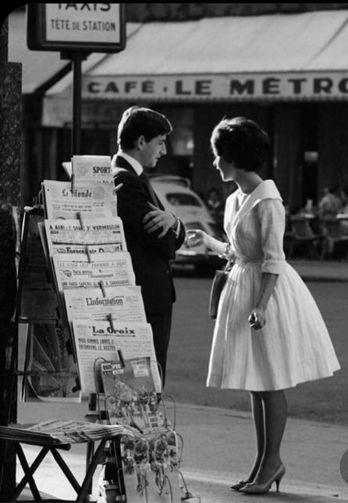 A black and white photo depicts a man and a woman conversing on a street next to a newsstand. The woman wears a knee-length dress, and the man is in a suit. Behind them is a café with a sign that reads "Le Métro." The atmosphere seems to be mid-20th century.