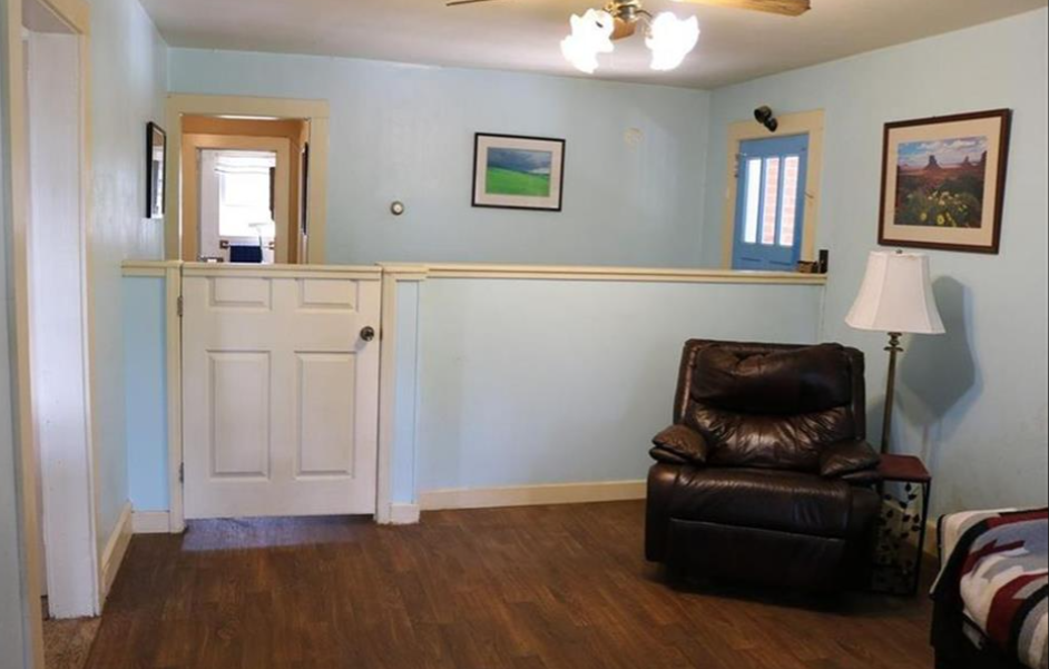 A cozy room features wooden flooring, light blue walls, and a brown leather recliner next to a white floor lamp. A half-door leads to an adjacent space. Framed pictures decorate the walls, and a ceiling light hangs above. A blue door is seen in the background.