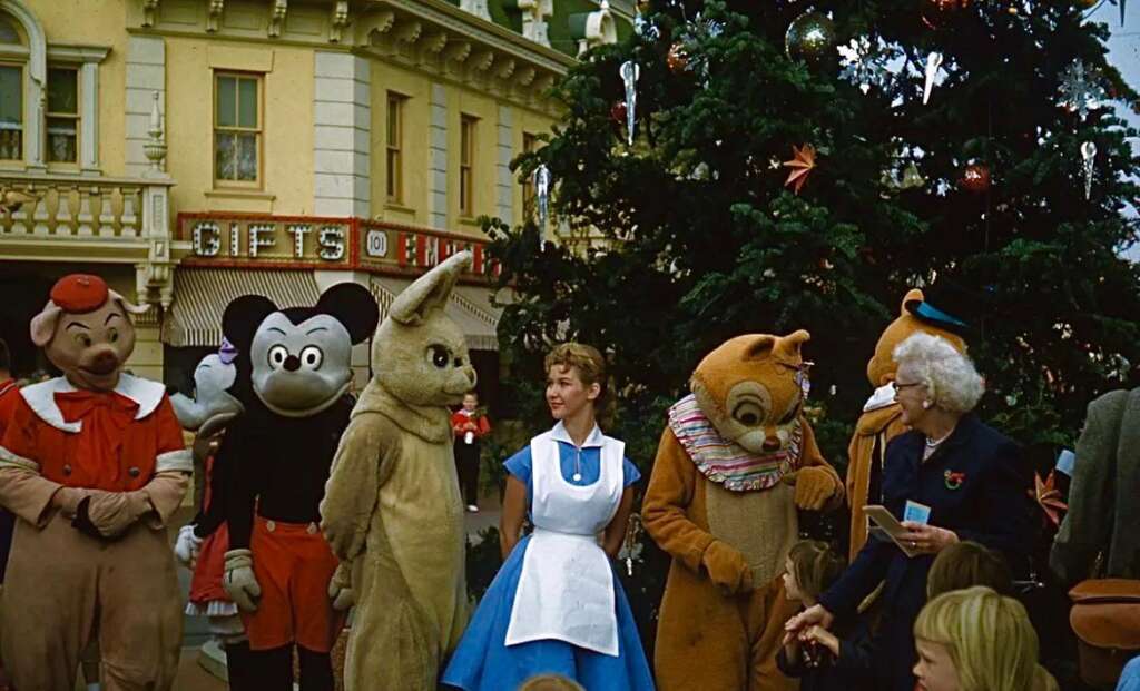 A group of people dressed in various character costumes, including Mickey Mouse, Chip, and others, pose in front of a large Christmas tree. They are outdoors near a building with a "GIFTS" sign. An older woman and another person in an Alice costume stand among them.