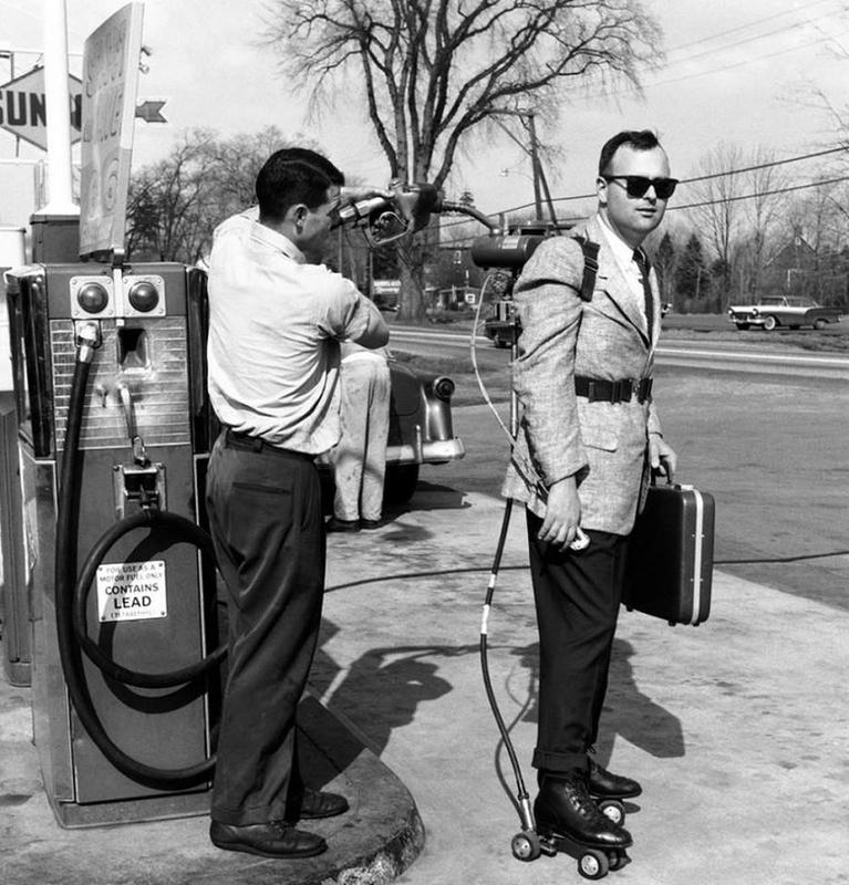 A man wearing roller skates, a jetpack, and a suit stands at a gas station. Another man is fueling the jetpack, which is connected to a gasoline pump. The scene appears to be from a mid-20th-century setting, with a vintage car visible in the background.