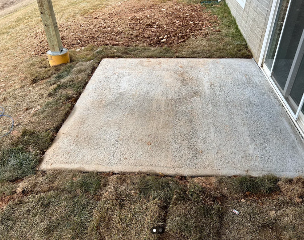 A small concrete patio slab is positioned next to a building with sliding glass doors. The surrounding area has patches of grass and exposed soil, indicating recent construction or landscaping work. A wooden post is seen embedded in the ground on the left side.