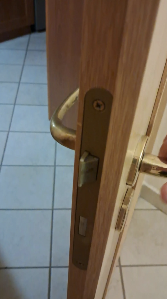 Close-up of a partially open wooden door with a brass handle. A person's hand is gripping the handle. The floor is tiled with white squares, and the background includes another door and beige wall.