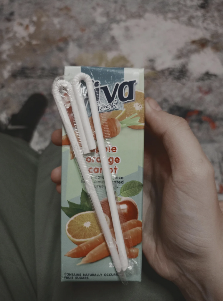 A person holds a Viva Fresh juice box with a visible label showing carrot and orange flavors. Two wrapped straws are attached to the front of the juice box. The background appears to be a mottled or painted surface.