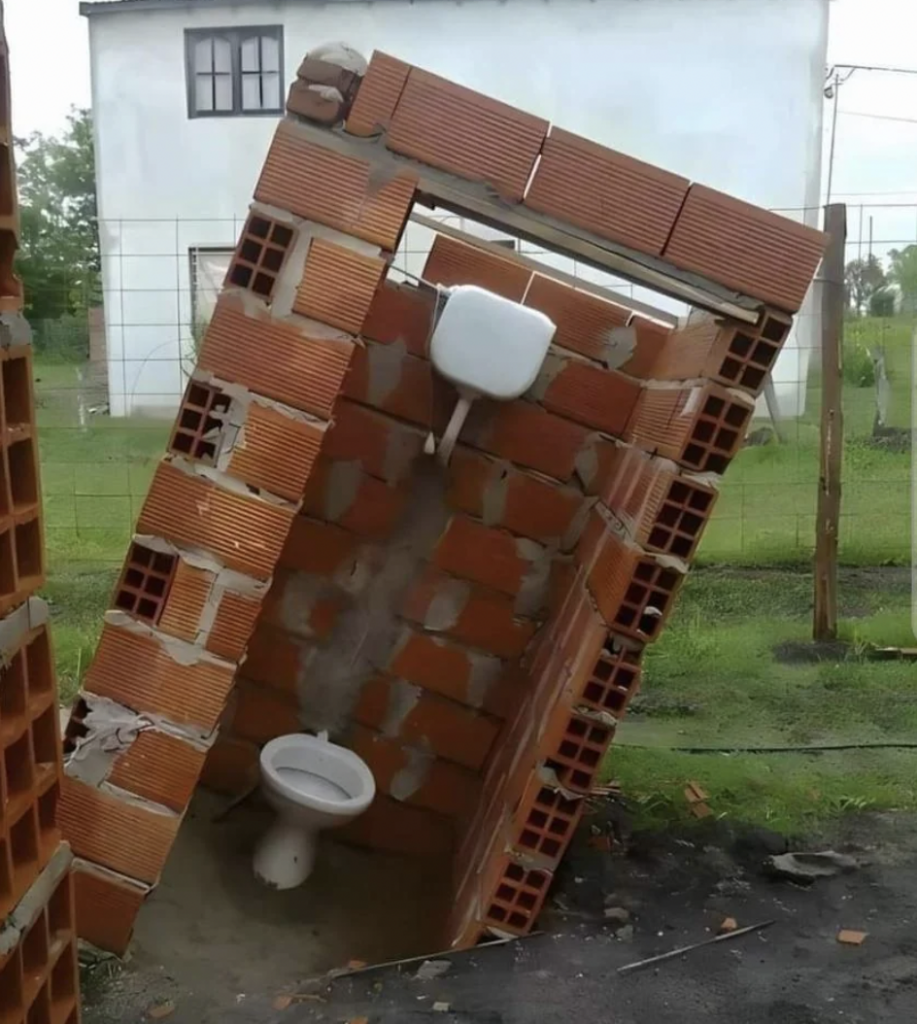 A small brick structure with a tiled roof and a toilet inside is partially collapsed and tilted sideways. The structure has holes in the walls and stands in a grassy outdoor area with a wire fence and a white building in the background.