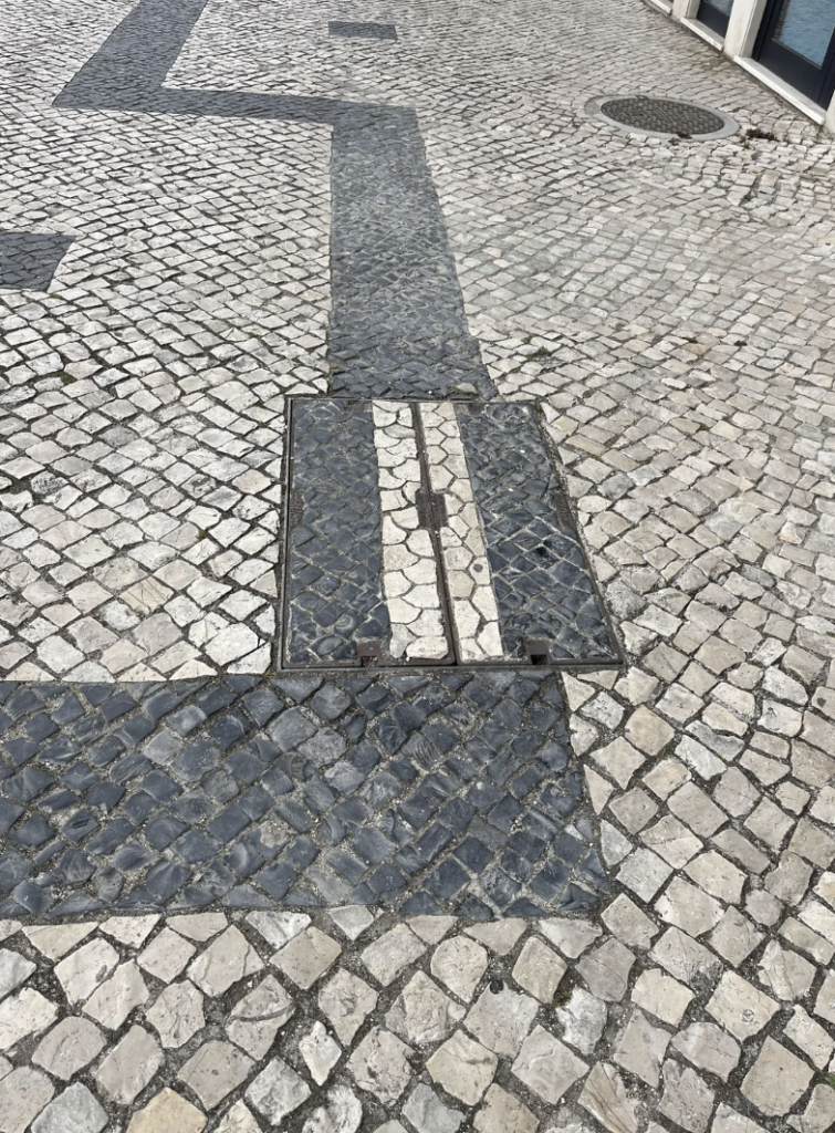 A cobblestone street with a rectangular manhole cover in the center. The manhole cover is aligned with the street's design and has dark and light stones that match the surrounding pattern. The stones around are predominantly light-colored, with some darker areas.