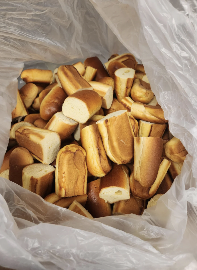 A large plastic bag filled with numerous pieces of sliced bread rolls. The bread appears to be lightly browned on the outside with a soft, white interior. The plastic bag is partially open to display the contents.