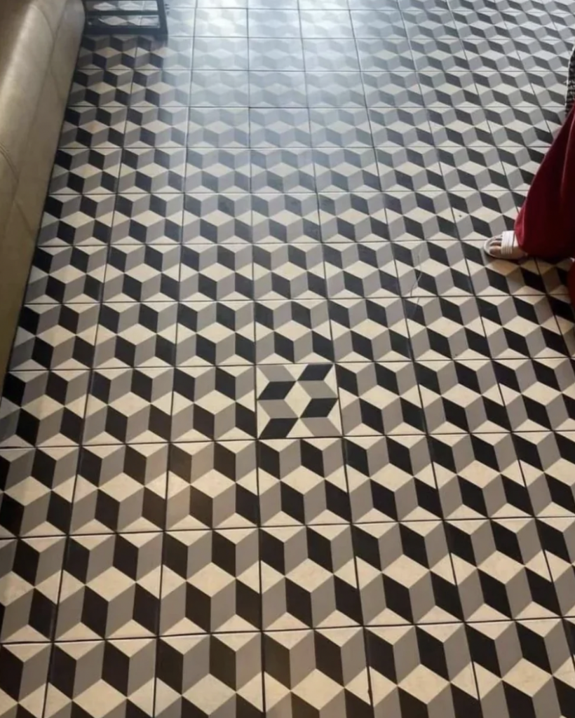 Geometric patterned tiled floor creating a 3D cube illusion in shades of white, gray, and black. Partially visible is a beige sofa, a small portion of a red garment, and a person's white sandal on the right side of the image.