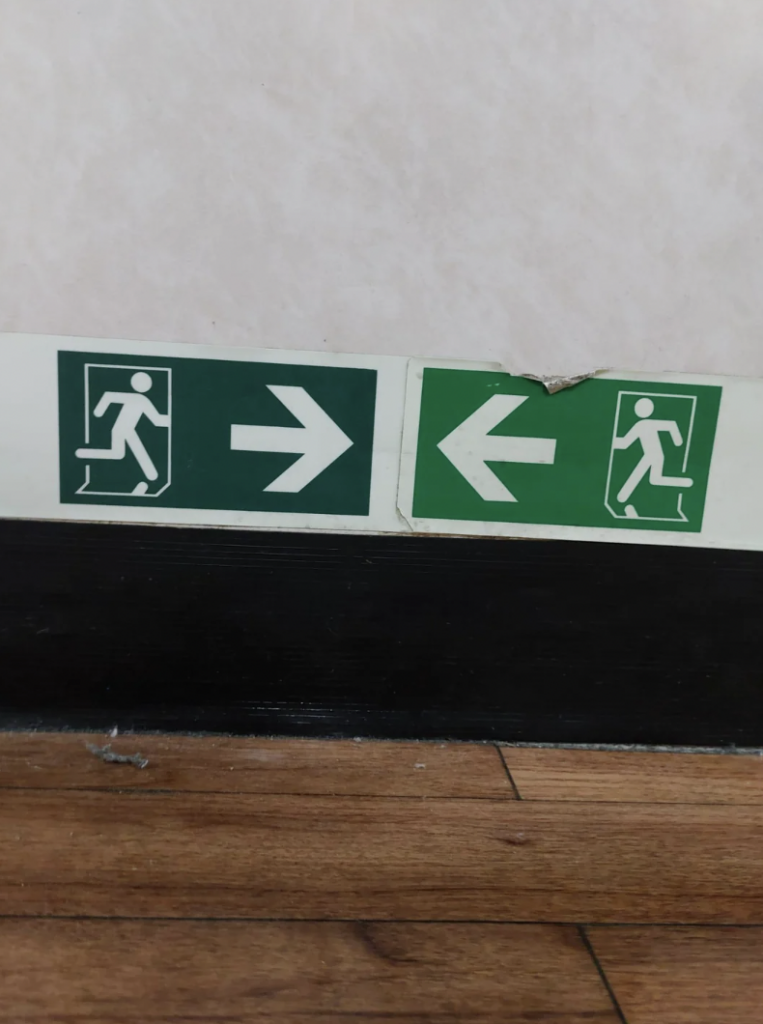 Two green emergency exit signs with white pictograms and arrows are mounted side by side on a wall. The left sign points right, and the right sign points left. The wall above has slight damage, and a wooden floor is visible at the bottom of the image.