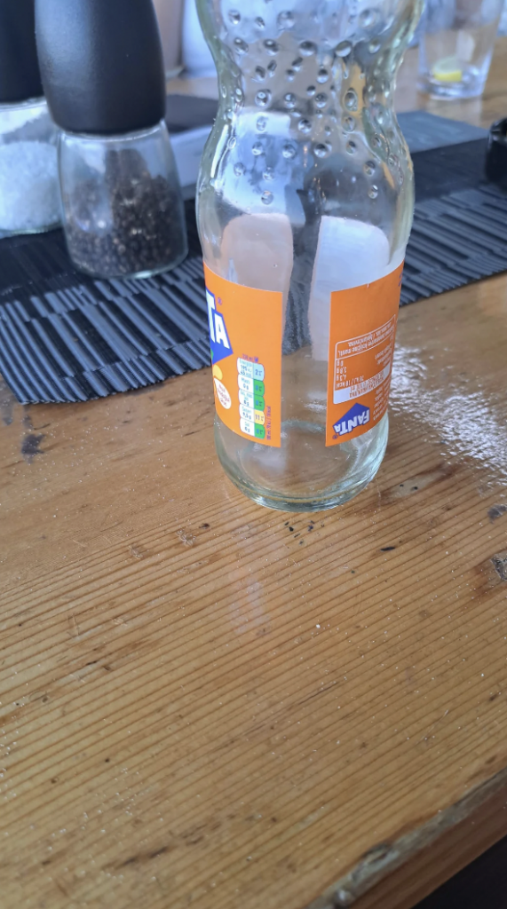 A glass bottle with an orange Fanta label sits empty on a wooden table. There is a clear bottle in the background, slightly out of focus. The table surface shows some water droplets and reflections. There is also a black placemat under the items.