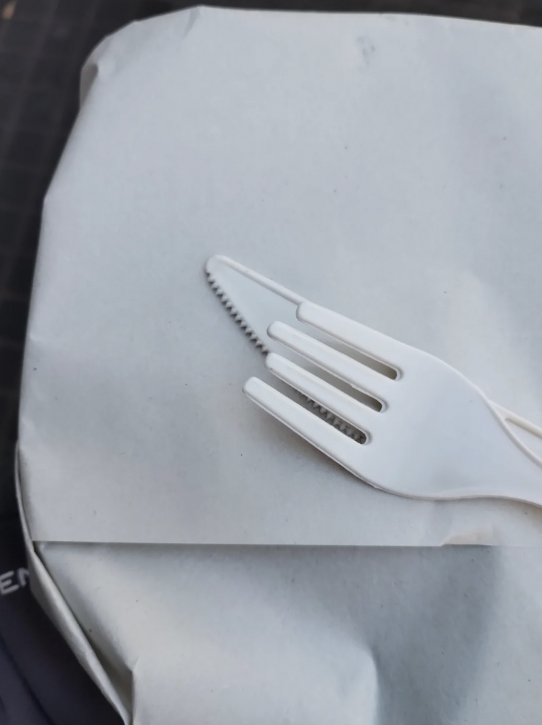 A white plastic fork with a serrated edge is placed on top of a white paper-wrapped item. The fork appears to also function as a knife. The background includes an indistinct surface with a dark element partially visible.