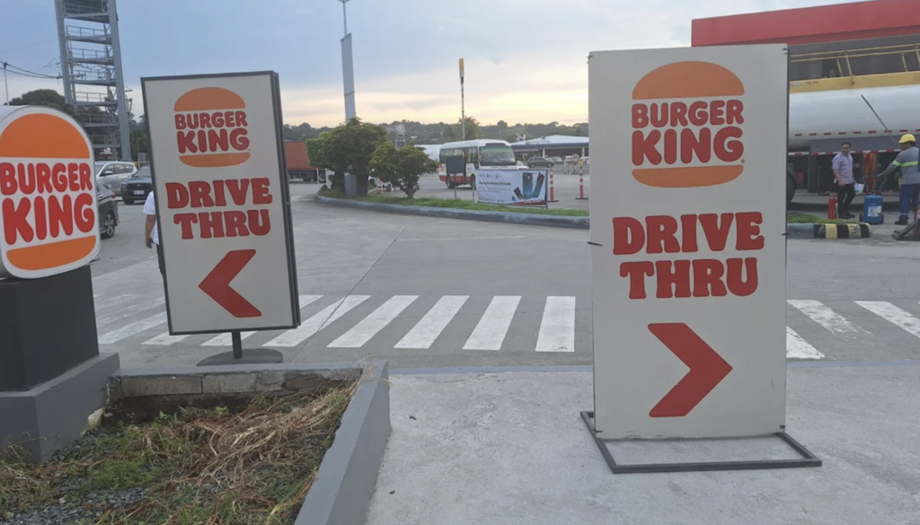 Parking area of a Burger King with drive-thru signs featuring arrows pointing to the drive-thru lane. Crosswalk and some construction activity visible in the background.