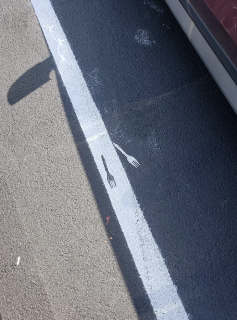 A fork is laying on a sunlit asphalt parking lot, partially casting a shadow while the other half appears painted, blending with the white line marking the parking space boundary. The shadow of a nearby vehicle is visible in the upper right part of the image.