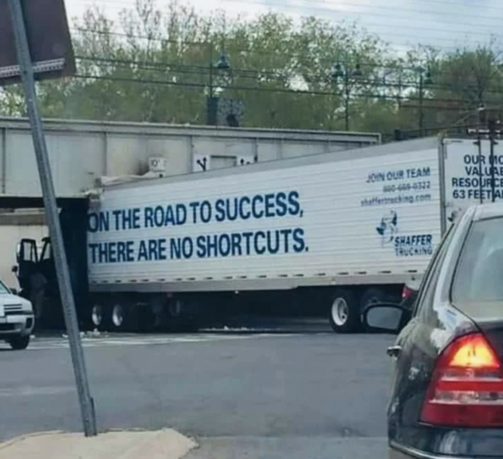 A large Shaffer Trucking semi-truck is stuck under a low bridge. The side of the truck trailer has the text "ON THE ROAD TO SUCCESS, THERE ARE NO SHORTCUTS," along with contact information for the trucking company.