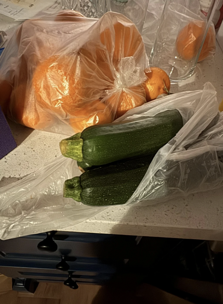 A plastic bag containing oranges is placed next to another plastic bag with two zucchinis on a kitchen countertop. In the background, there are glasses and other kitchen items. The countertop appears cluttered.