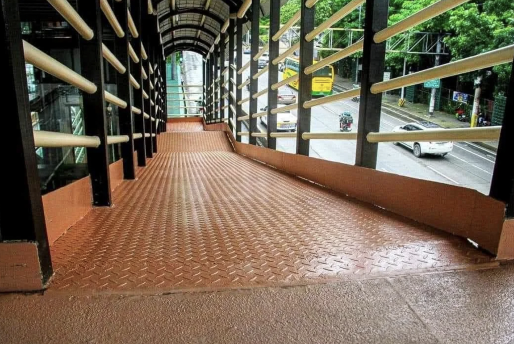 An inclined pedestrian pathway made of textured brown metal with angled handrails and a covered roof. The pathway leads down towards a bustling street with trees and vehicles visible in the background.