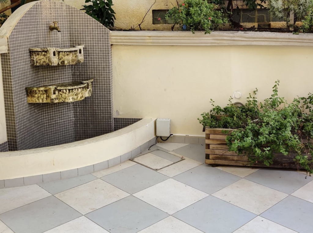 A tiled courtyard with an old, rustic wall fountain featuring two basins. To the right, a large wooden planter box holds a variety of green plants. The area is lined with beige and gray square tiles. An electrical box is mounted on the wall near the planter.