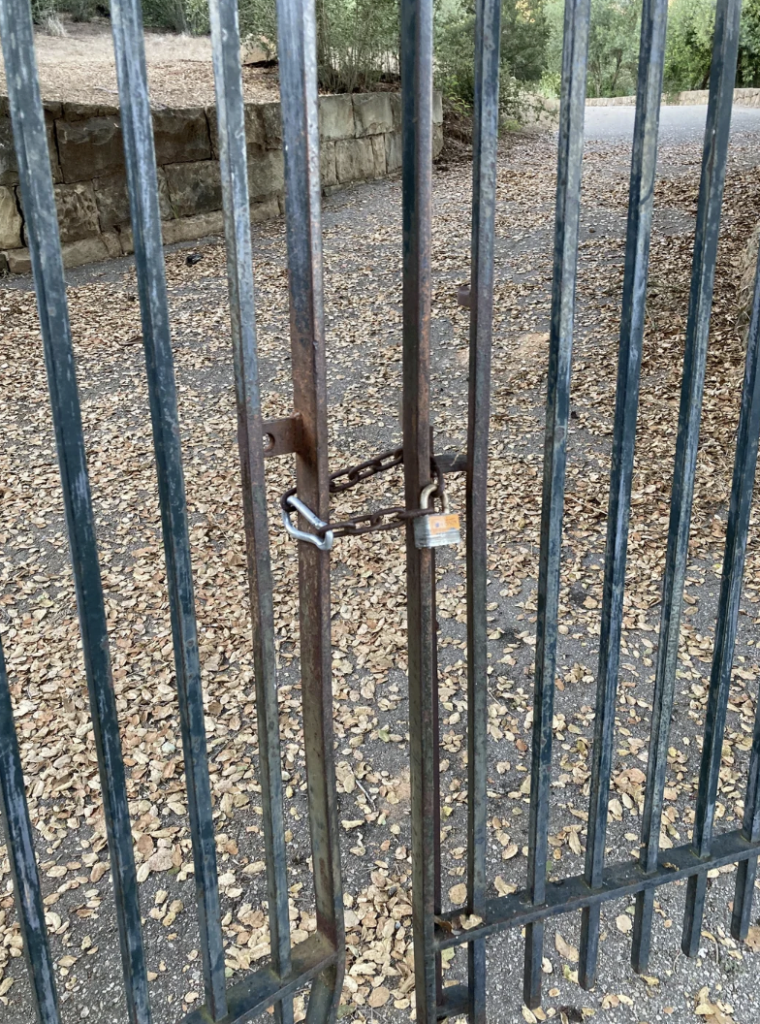 A close-up view of a locked metal gate with vertical bars. The gate is secured with two padlocks. There are dry leaves scattered on the ground in front of the gate, and a stone wall is visible in the background. Trees and a pathway can be seen in the distance.